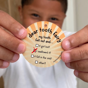 a boy holds a lost tooth token that says Dear Tooth Fairy my tooth fell out and... it got lost, I swallowed it, it fell in the sink, other. 